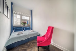 A bedroom with a bed, a window, a chair, and a view of a building outside. - Apartment Żelazna 58 (Warszawa)