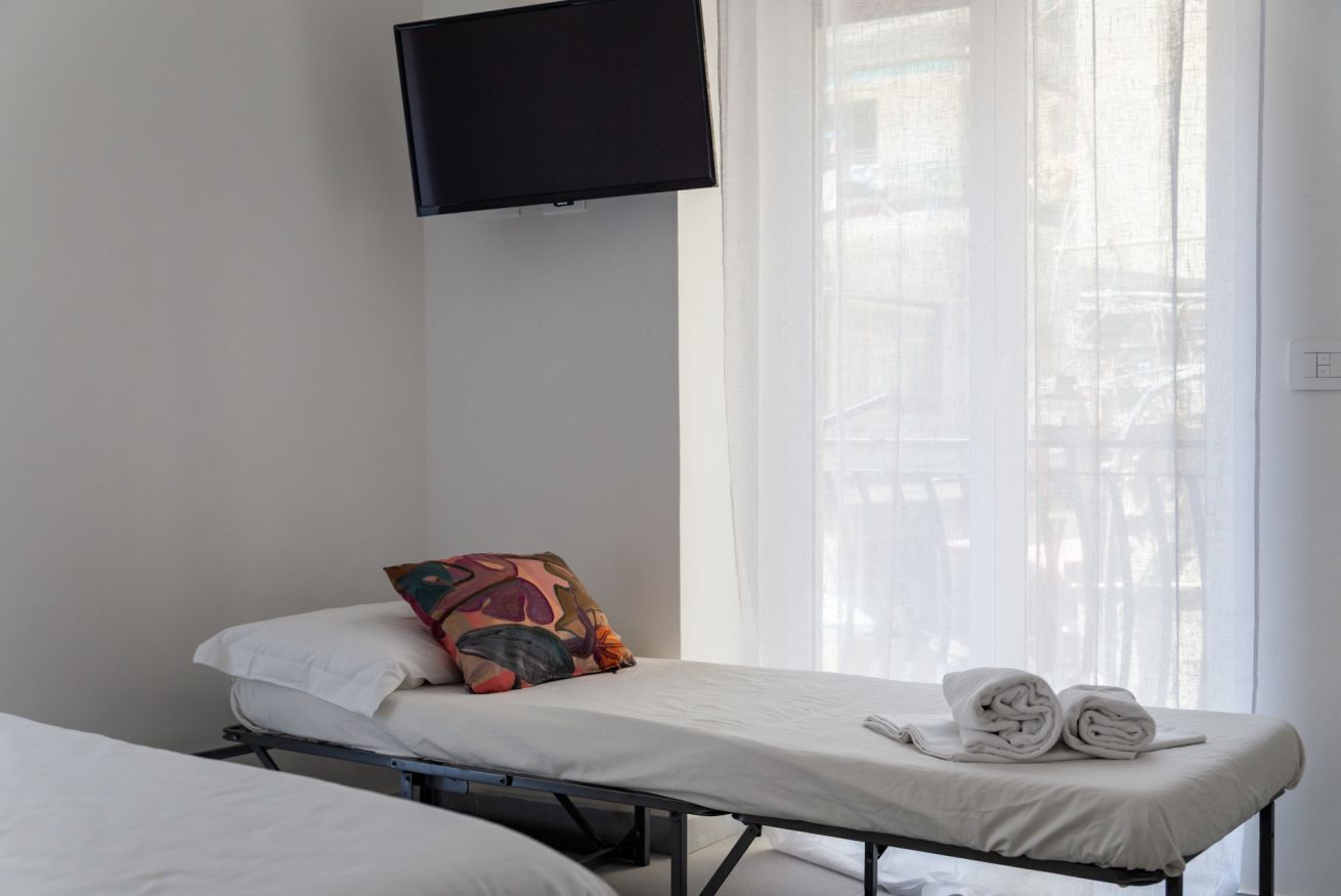 Ein Einzelbett mit weißer Matratze und einem bunten Kissen, ein aufgerolltes Handtuch und ein an der Wand befestigter Fernseher.