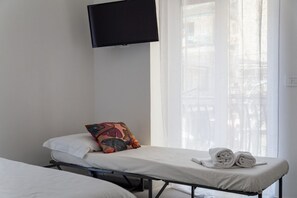 Een eenpersoonsbed met een witte matras en een kleurrijk kussen, een opgerolde handdoek en een aan de muur bevestigde televisie.