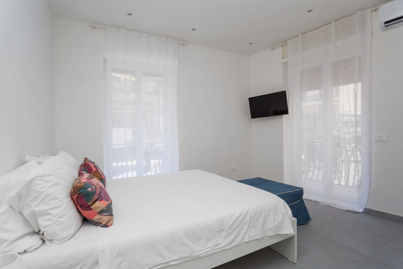 Ein Schlafzimmer mit einem Bett, einem an der Wand befestigten Fernseher und einem Fenster mit durchsichtigen Vorhängen.