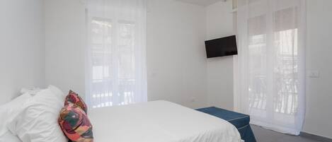 Une chambre à coucher avec un lit, un téléviseur fixé au mur et une fenêtre ornée de rideaux légers.
