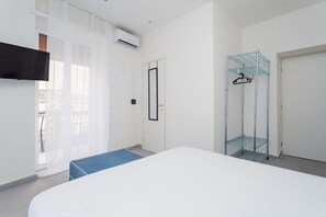 Een slaapkamer met een bed, een aan de muur bevestigde televisie, een airconditioningapparaat, een deur en een glazen open kast met kledinghangers.