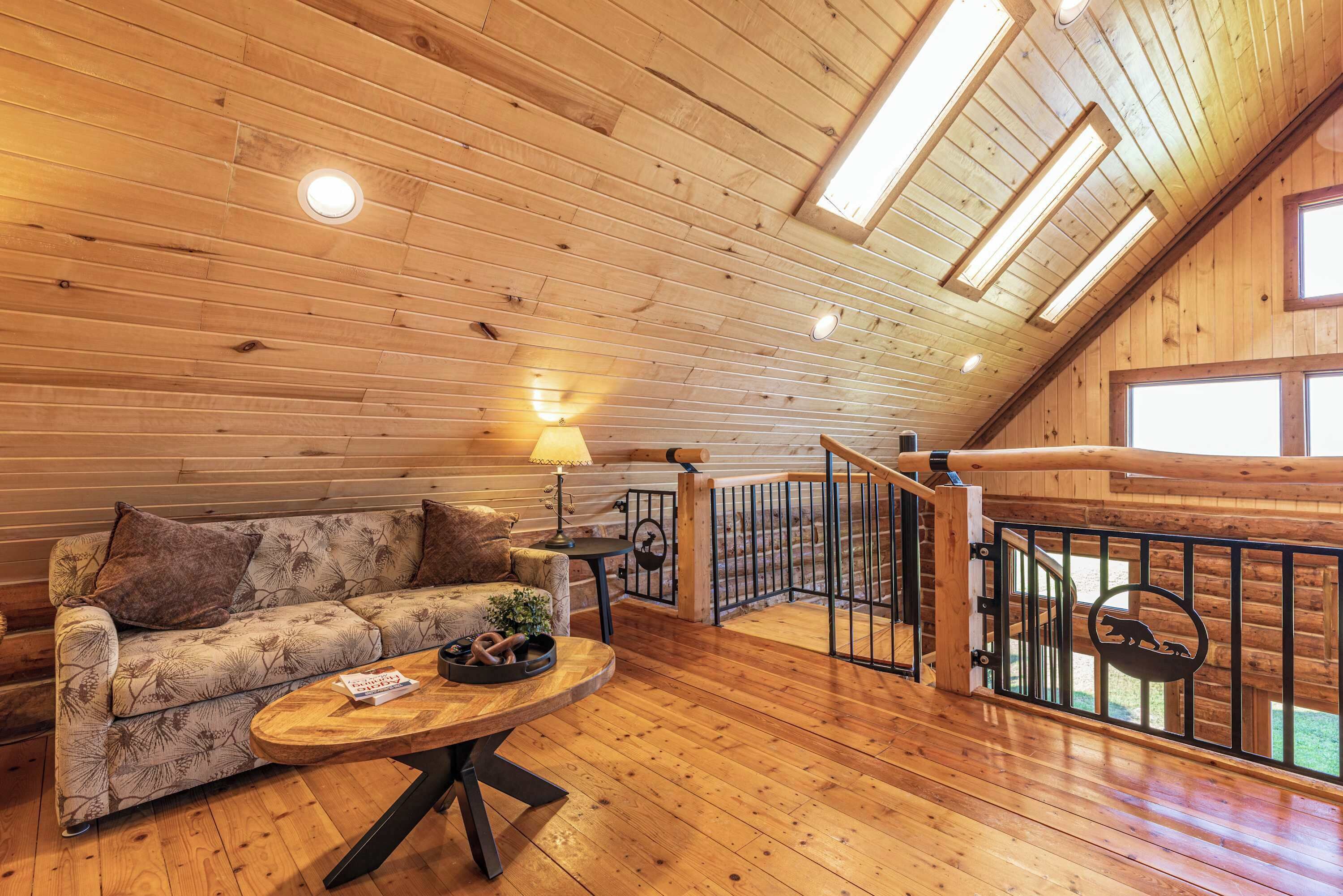 A cozy cabin interior with a wooden ceiling, a sofa, a round wooden table, and a staircase with a railing.