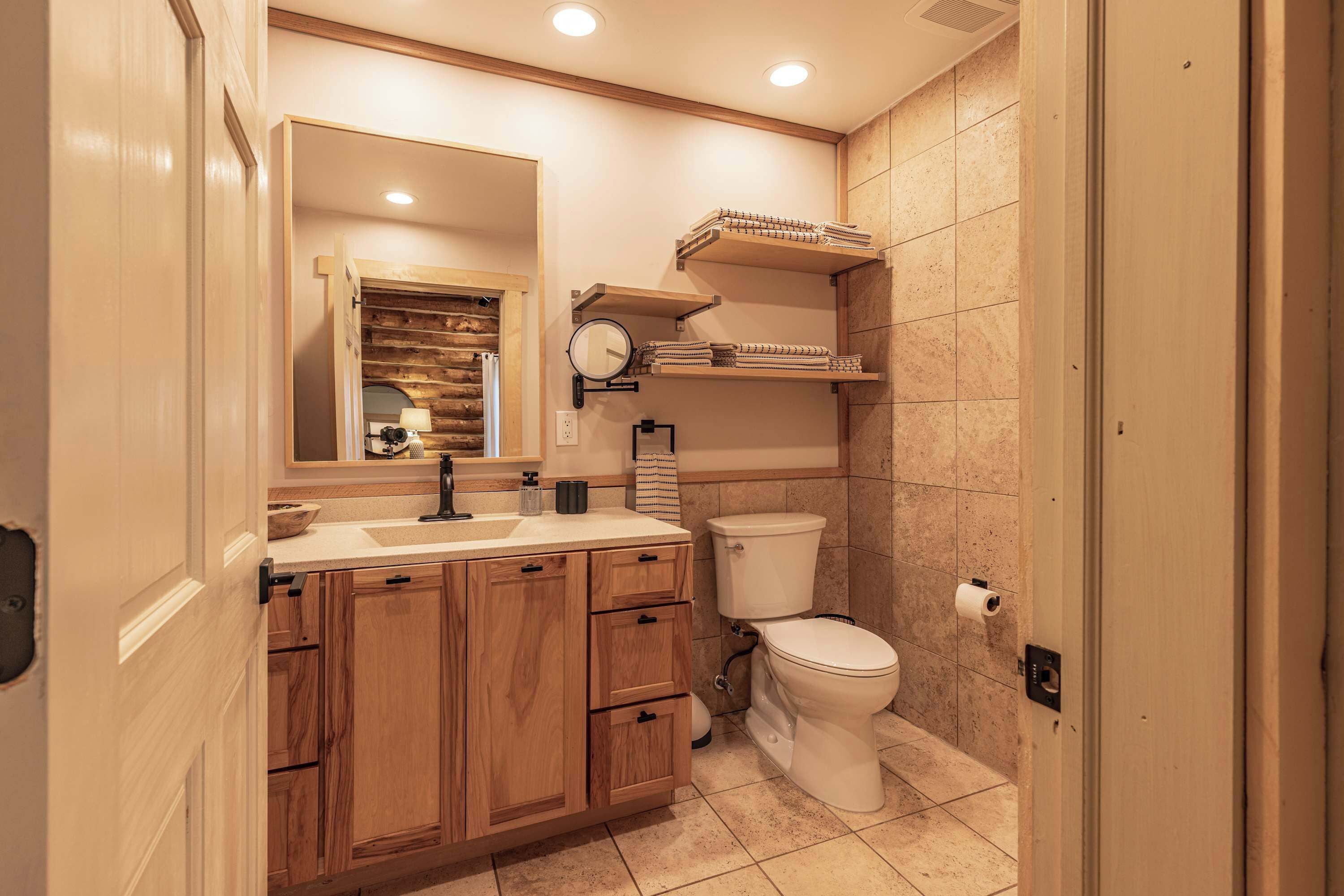 A bathroom with wooden cabinets, a sink, a mirror, a toilet, and shelves above the toilet.