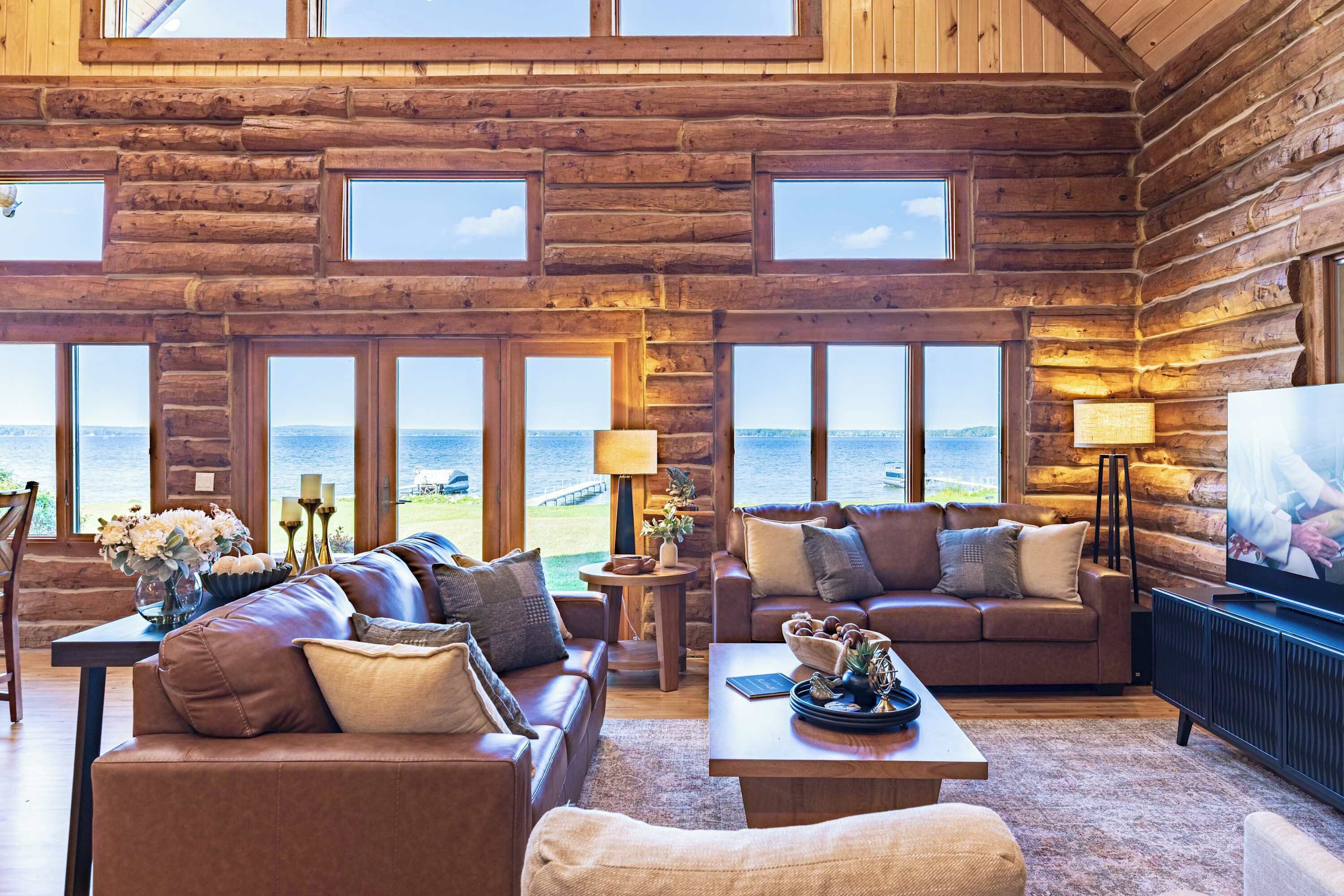 Interior de cabana de tronco com grande janela com vista para um corpo de água, mobiliada com sofás de couro e uma televisão.