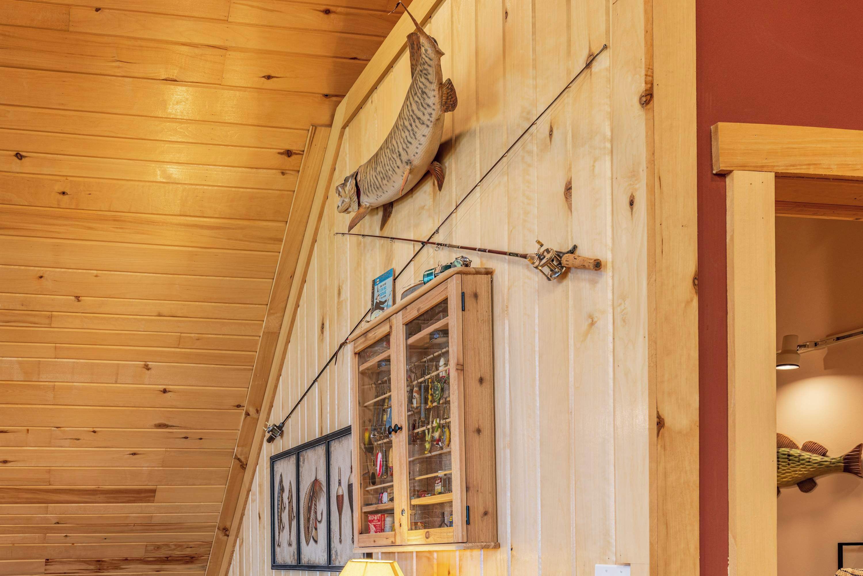 A wooden interior with a fishing rod mounted on the wall, a window with fishing lures, and a lamp.