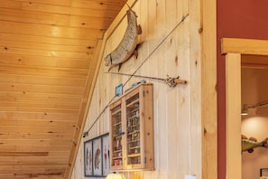 A wooden interior with a fishing rod mounted on the wall, a window with fishing lures, and a lamp.
