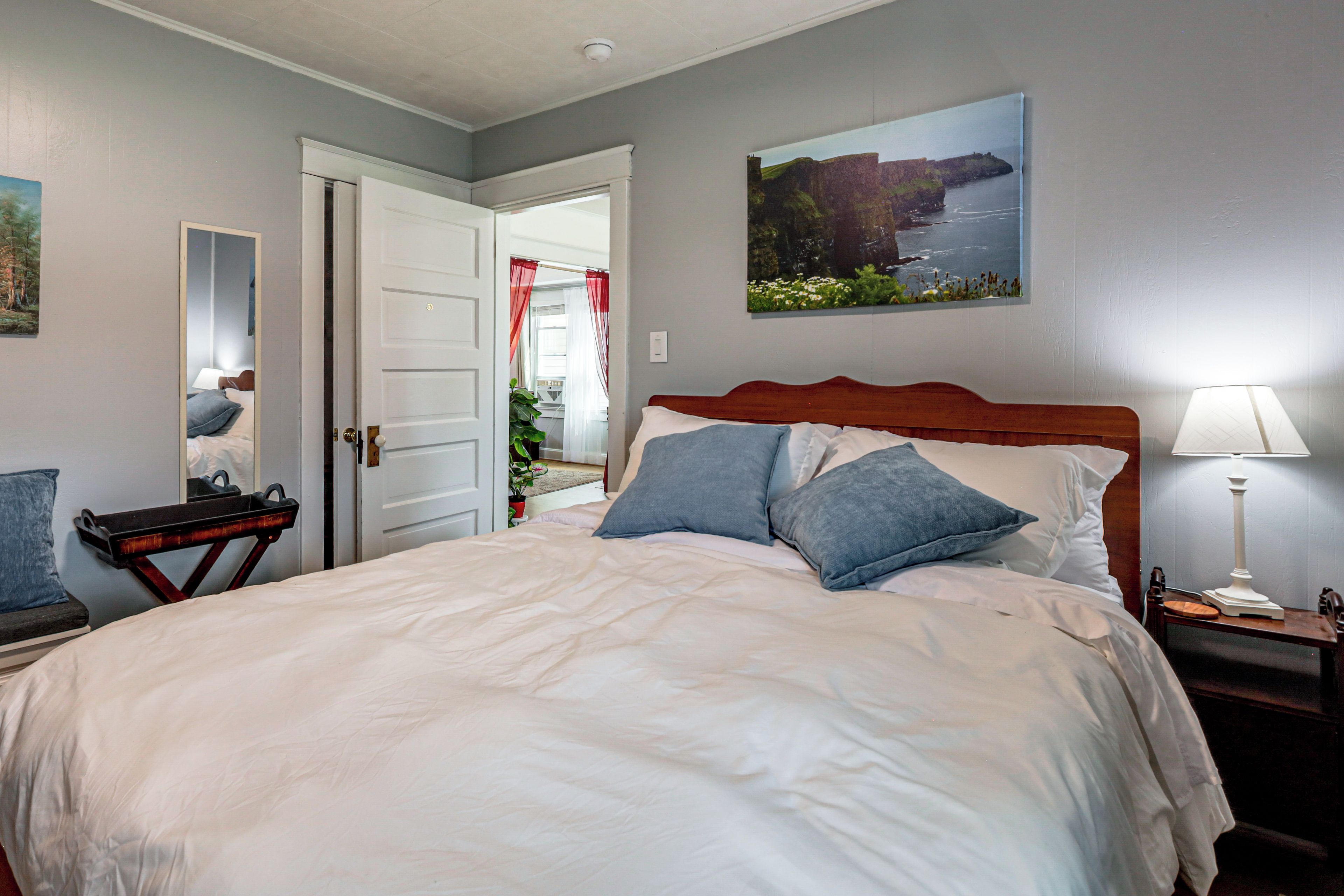 A bedroom with a large bed, a bedside table with a lamp, a chair, and a door leading to another room.