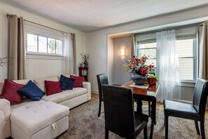 A living room with a white sofa, blue and red cushions, a dining table with chairs, and a vase of flowers.