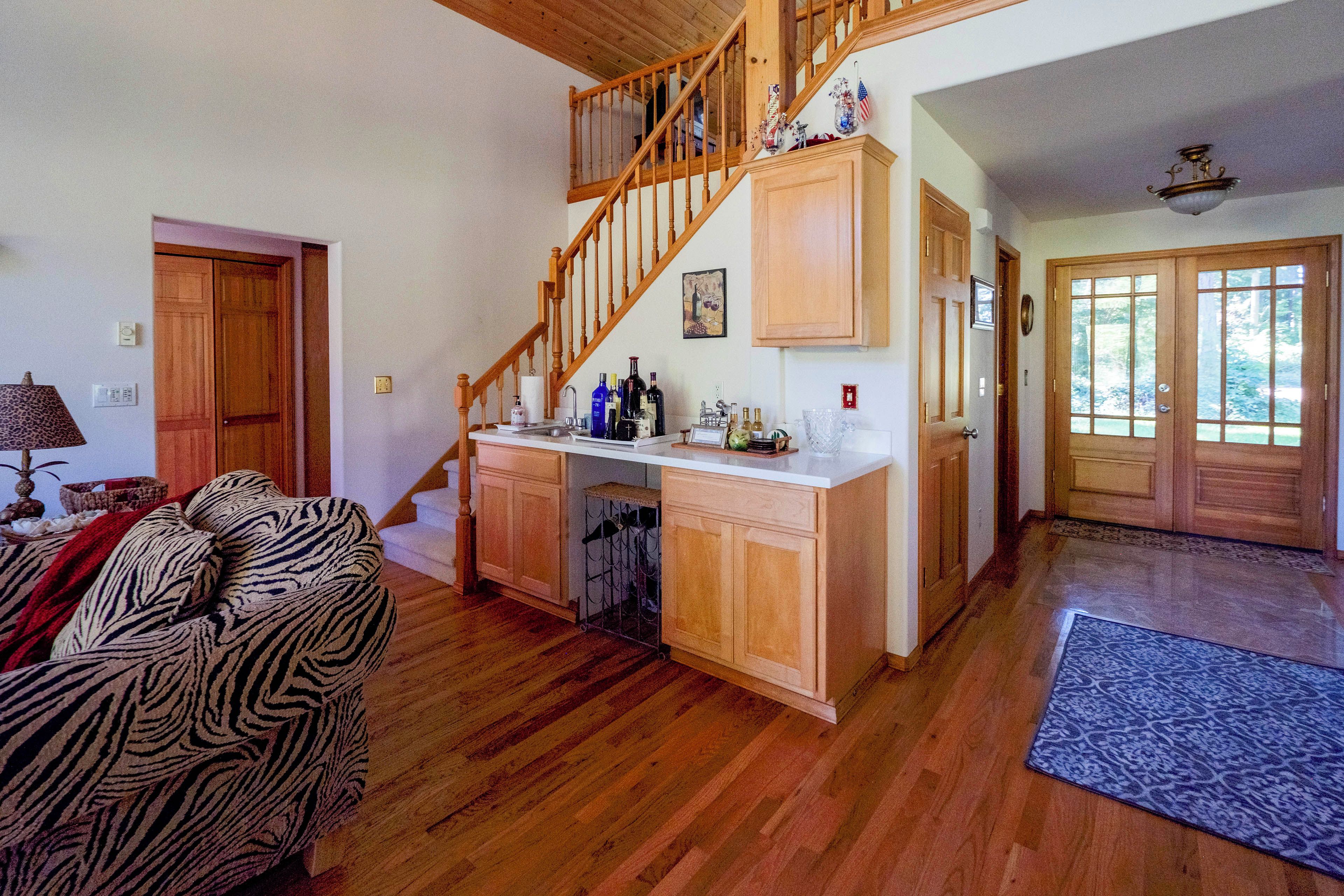 A spacious living area with a wooden staircase, a kitchen with a white countertop, and a view of the outdoors through a door.