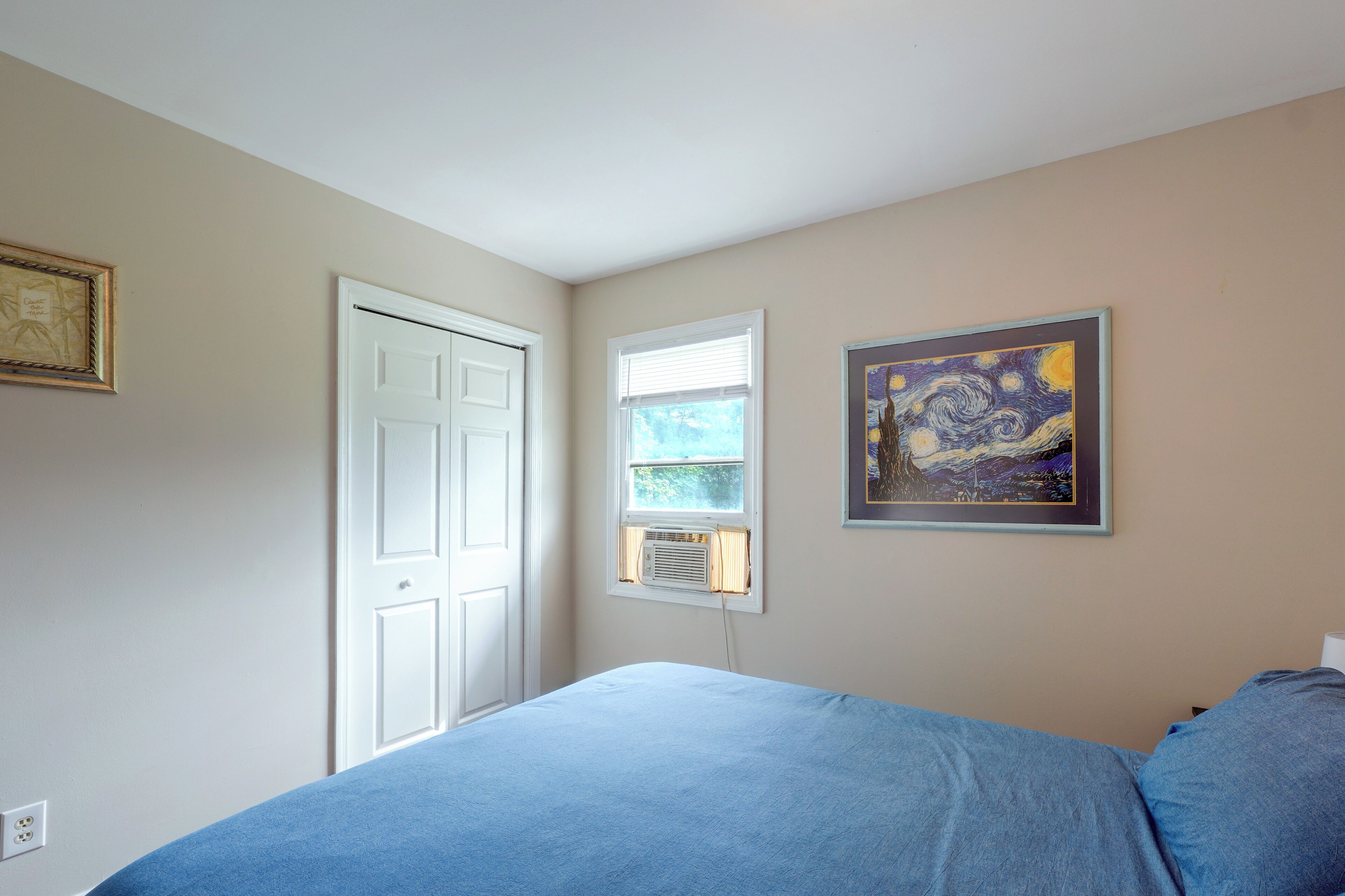 A bedroom with a bed, a door, a window, and two framed artworks on the wall.