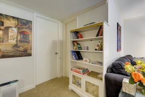 A room with a white bookshelf containing books and board games, a painting on the wall, and a grey sofa with a vase of flowers. - Easy Raleigh Access! Quaint Apt w/ Patio (Garner)