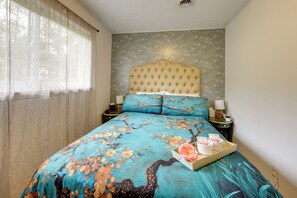 A bedroom with a floral bedspread, a tufted headboard, and a bedside table with a lamp and a tray of items. - Easy Raleigh Access! Quaint Apt w/ Patio (Garner)