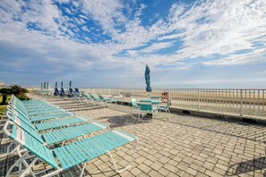 Een terras aan het strand met rijen blauwe ligbedden en enkele tafels, met uitzicht op het strand.
