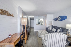 A cozy living room with a sofa, ottoman, and a dining area with a wooden table and chairs. - Steps to the Sand: Chic Daytona Oceanfront Condo (Daytona Beach)