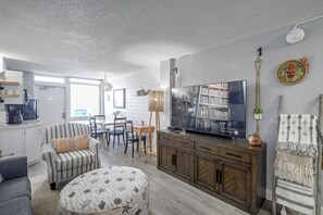 Un salon moderne doté d’un téléviseur à écran plat, d’un canapé à rayures, d’un pouf rond et d’un coin repas avec des chaises et une table.