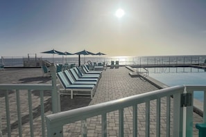 A sunlit outdoor seating area with lounge chairs and umbrellas, overlooking a calm body of water. - Steps to the Sand: Chic Daytona Oceanfront Condo (Daytona Beach)