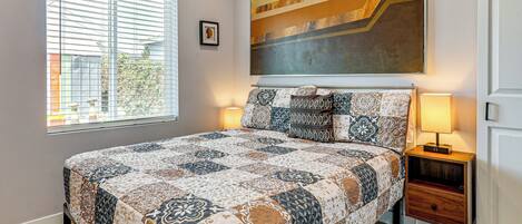 A bedroom with a large bed, a nightstand, a lamp, a window with blinds, and a painting on the wall.