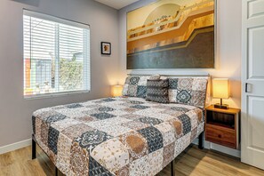 A bedroom with a large bed, a nightstand, a lamp, a window with blinds, and a painting on the wall.
