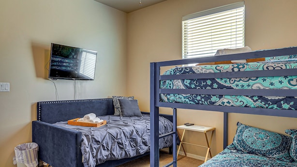 Une chambre avec un lit superposé, un lit simple, un téléviseur fixé au mur, une petite table et une fenêtre avec des stores.