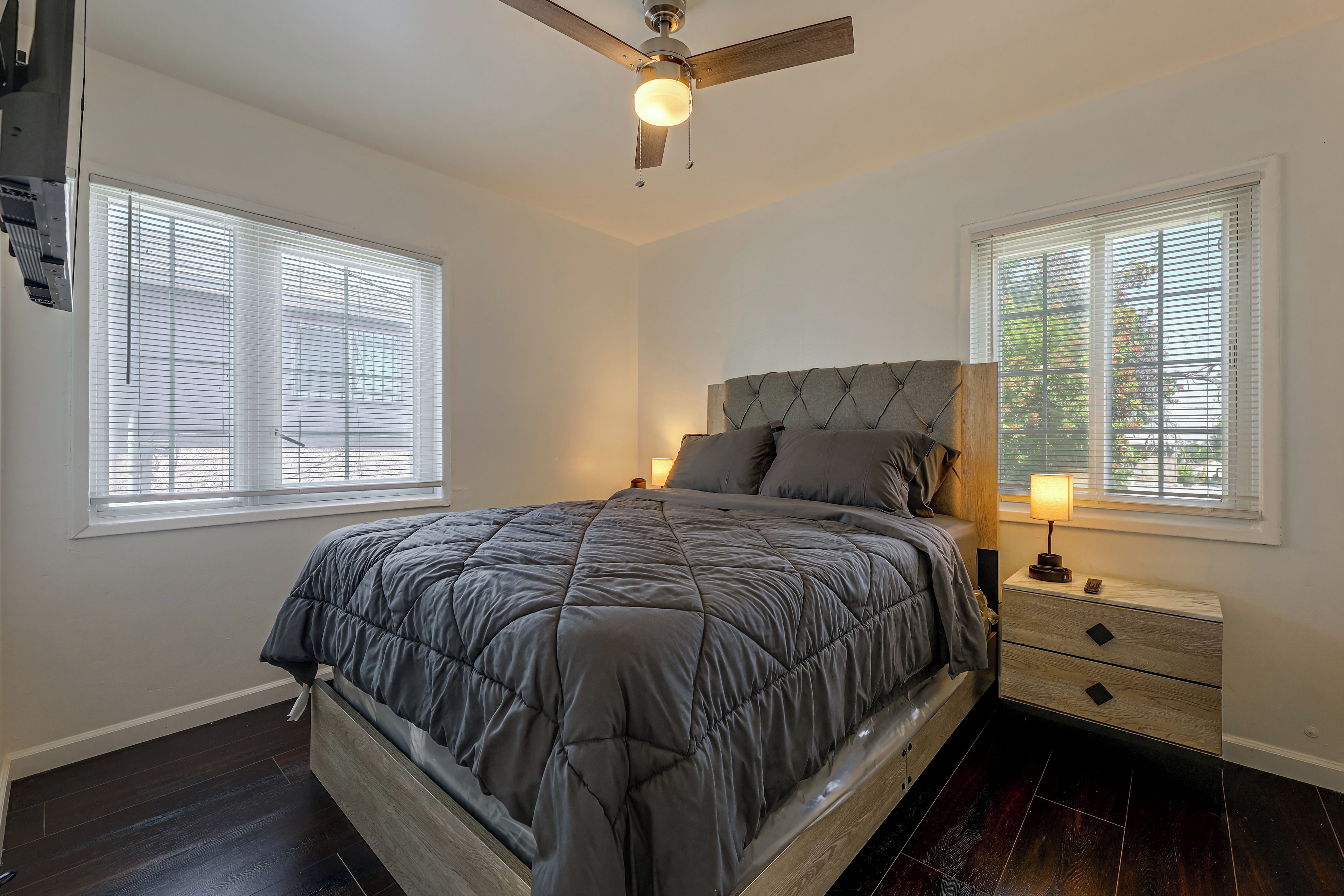A bedroom with a bed, a nightstand, a lamp, a ceiling fan, and windows with blinds.