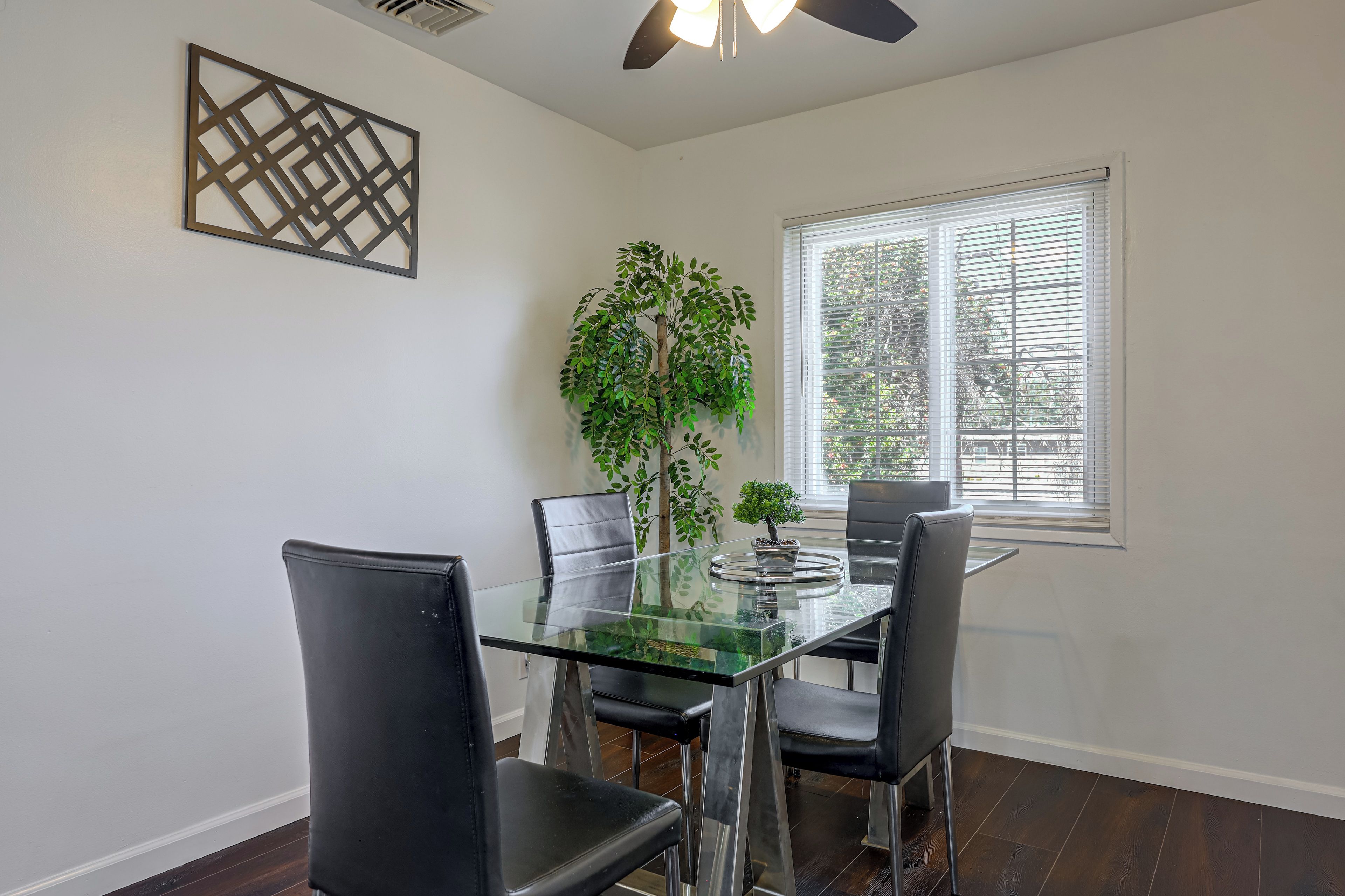 A dining area with a glass table, four chairs, a potted plant, and a window with blinds.
