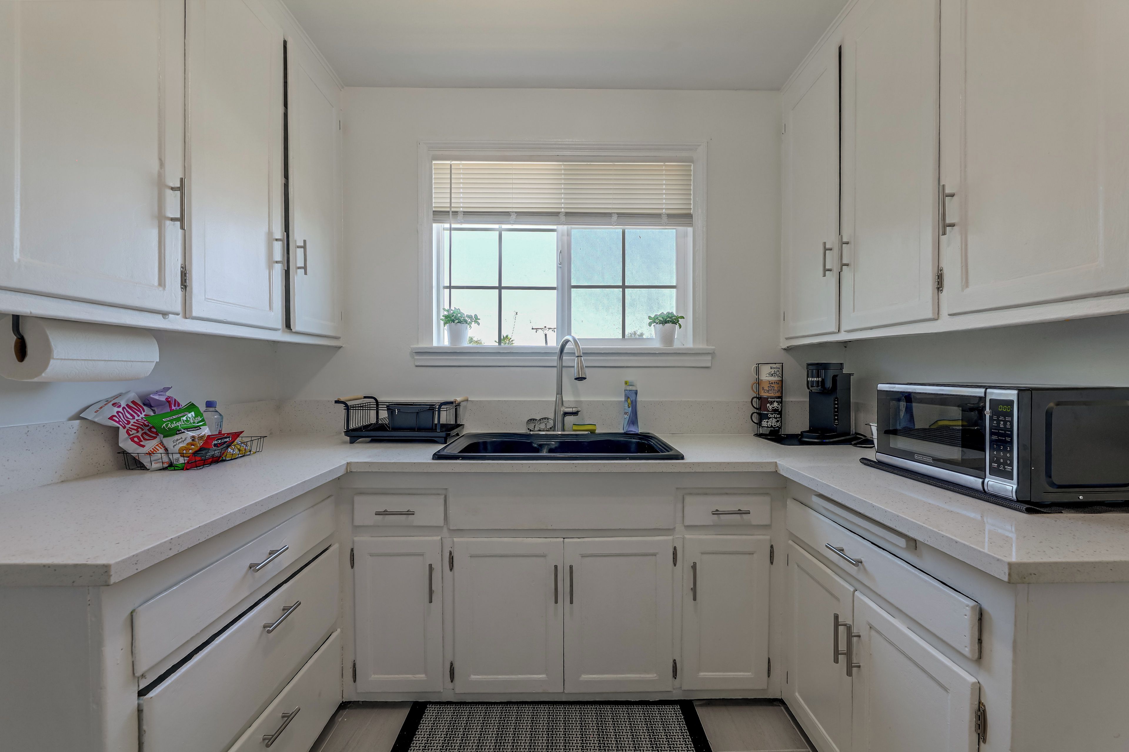 A kitchen with white cabinets, a window with blinds, a sink, a microwave, and a coffee maker.