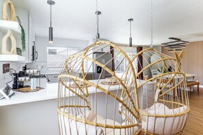 A modern kitchen with a coffee station, hanging light fixtures, and a dining area with a rattan chair.