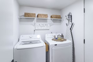 A laundry room with two white washing machines, a sign above them, and a shelf with baskets.