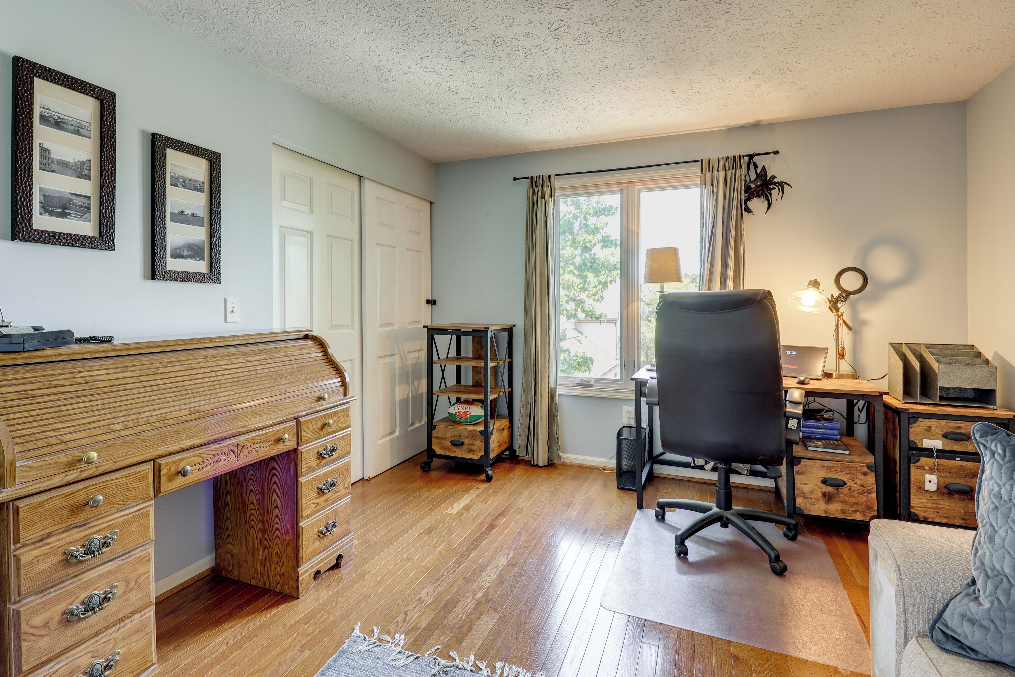 Une pièce avec un bureau en bois, un fauteuil de bureau noir, un bureau informatique avec une lampe et une fenêtre avec des rideaux.
