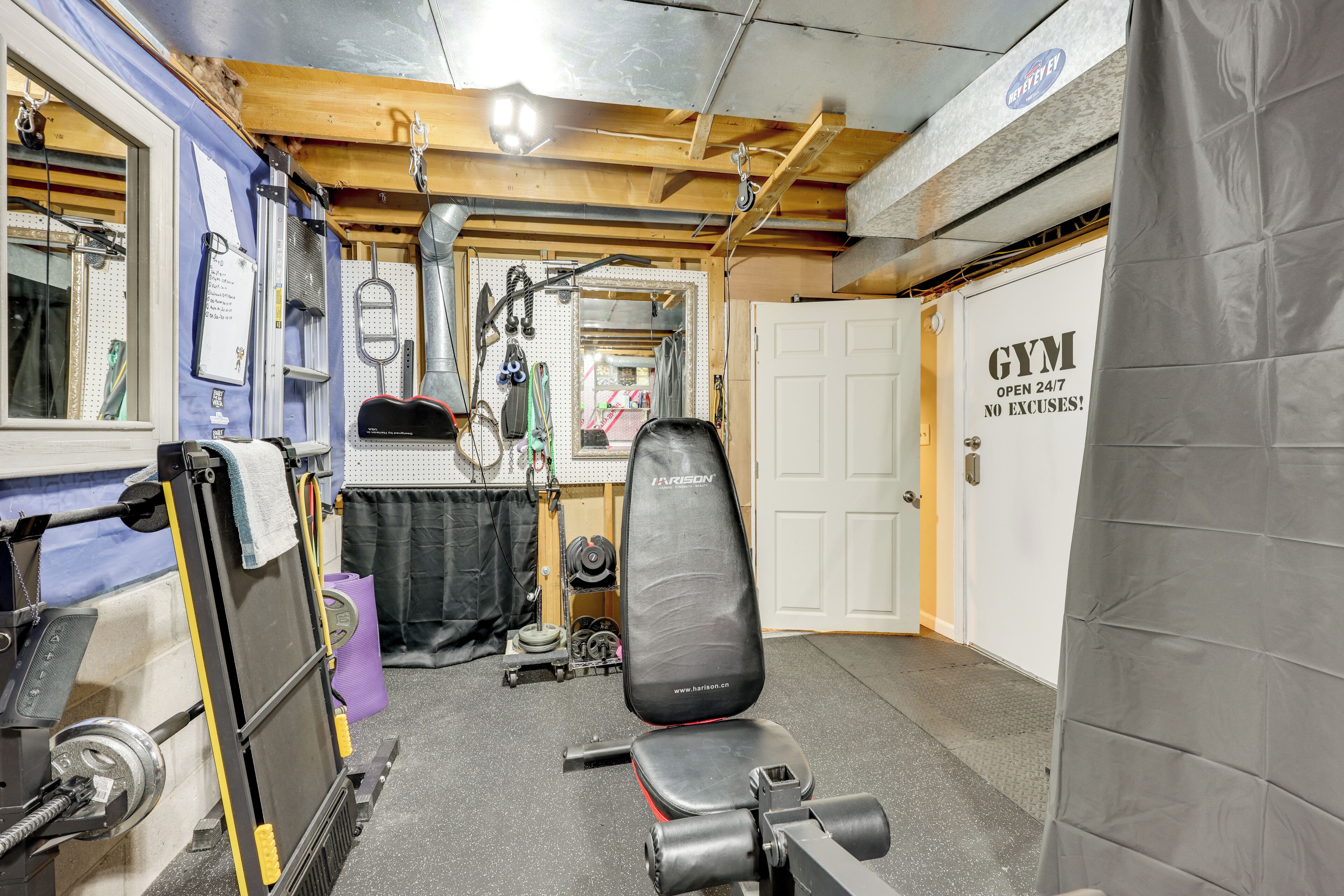 A gym with a weight bench, various exercise equipment, and a door labeled 'GYM OPEN 24/7 NO EXCUSES!'