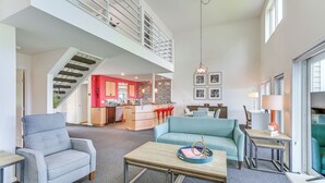 Een ruime woonkamer met een hoge plafond, een trap naar een hoger gelegen verdieping, een keuken met rode details en een eethoek met een blauwe bank.