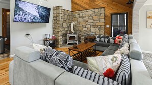 A modern living room with a stone wall, a fireplace, a large flat-screen TV, and a grey sectional sofa. - Group Getaway w/ Lake Views in Bronston! (Bronston)