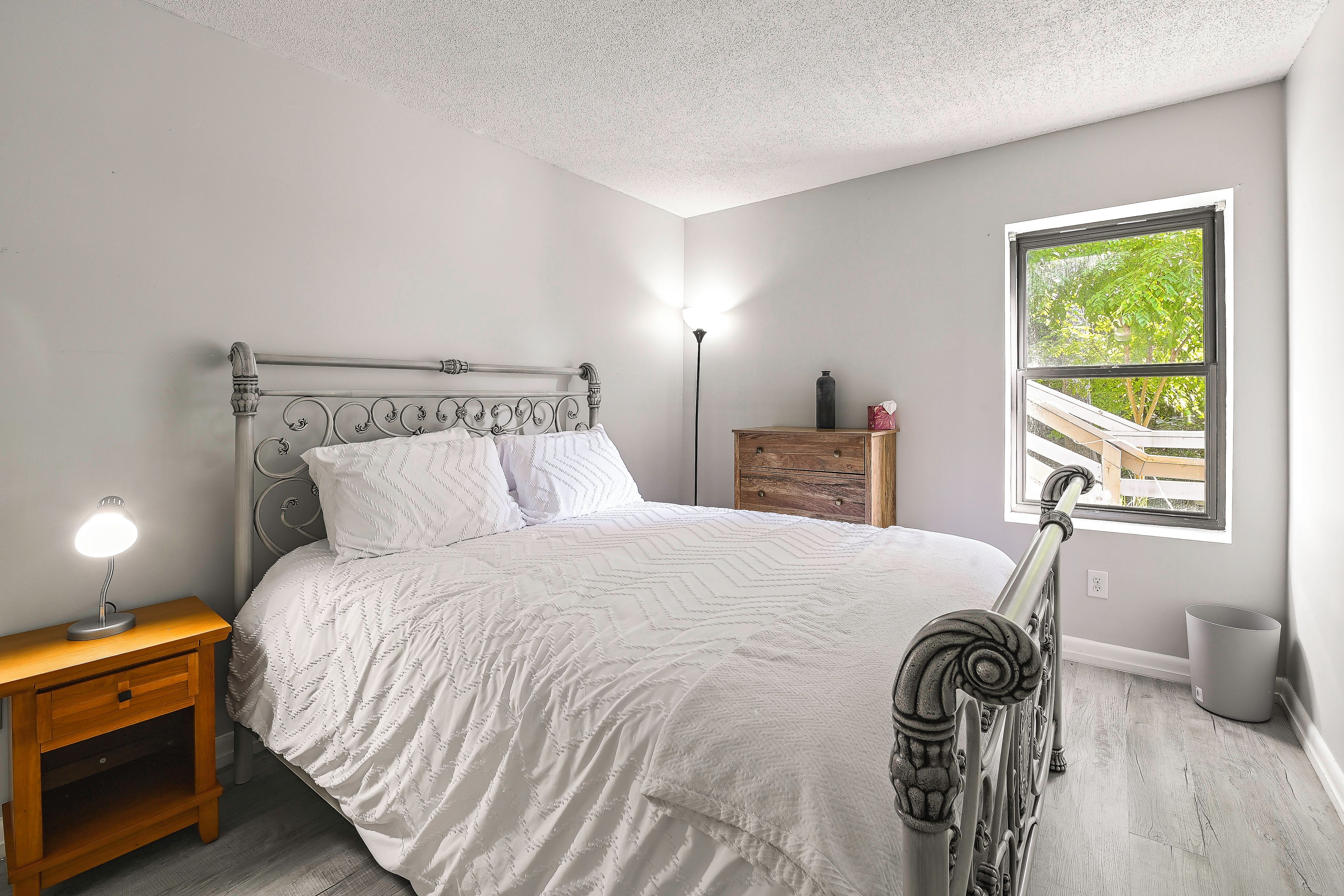 A bedroom with a bed, bedside table, lamp, dresser, and a window with a view.
