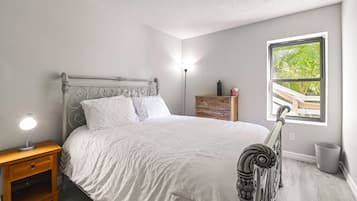A bedroom with a bed, bedside table, lamp, dresser, and a window with a view.