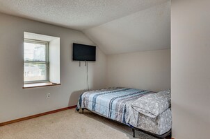 A bedroom with a single bed, a window with blinds, a flat-screen TV mounted on the wall, and a carpeted floor.