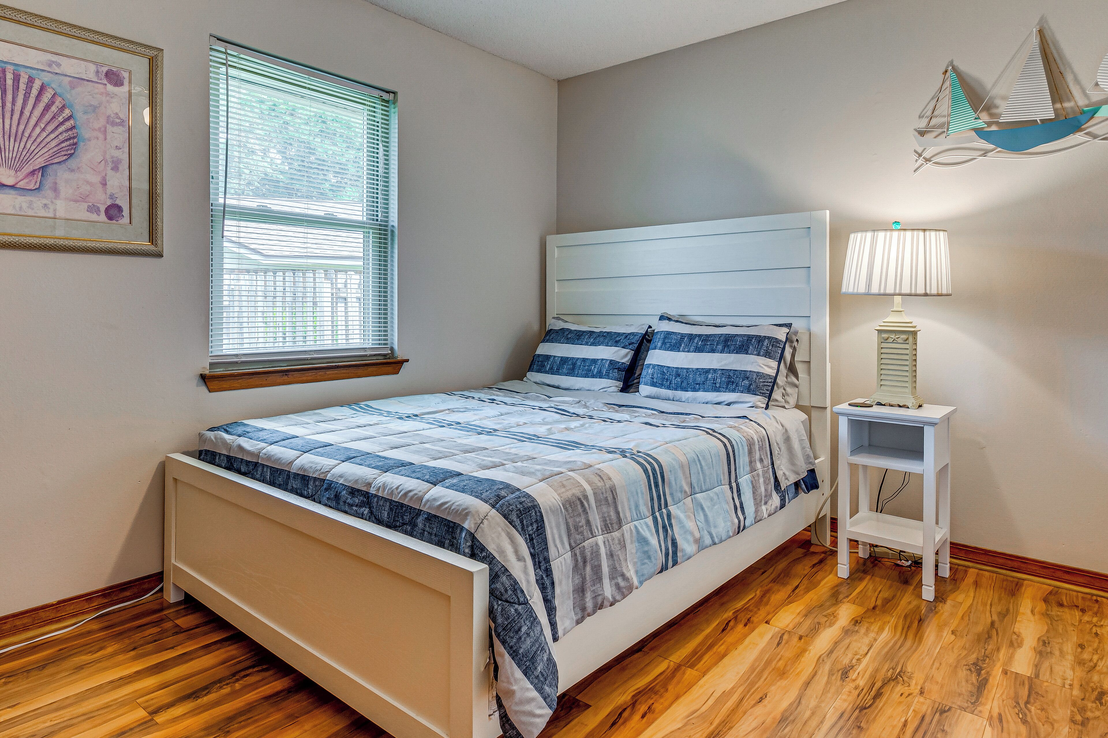 A bedroom with a bed, a nightstand, a lamp, a window with blinds, and a wall decoration.