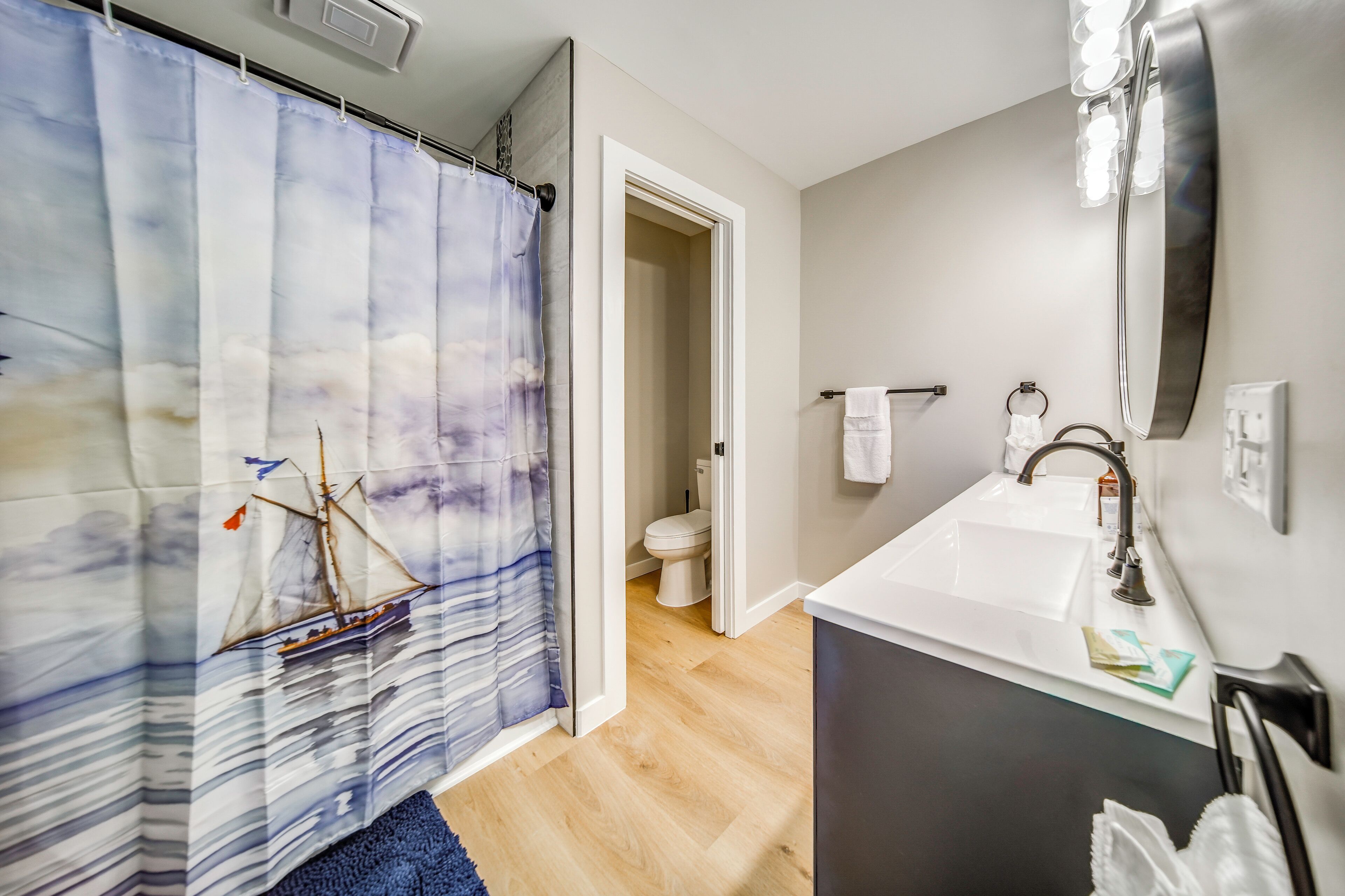 A bathroom with a shower curtain depicting a sailboat, a white sink with a mirror above it, and a toilet visible through an open door.