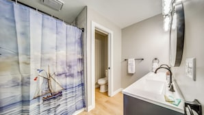 A bathroom with a shower curtain depicting a sailboat, a white sink with a mirror above it, and a toilet visible through an open door. - Family Fun! Indoor Jungle Gym, Hot Tub & Beach (Erie)