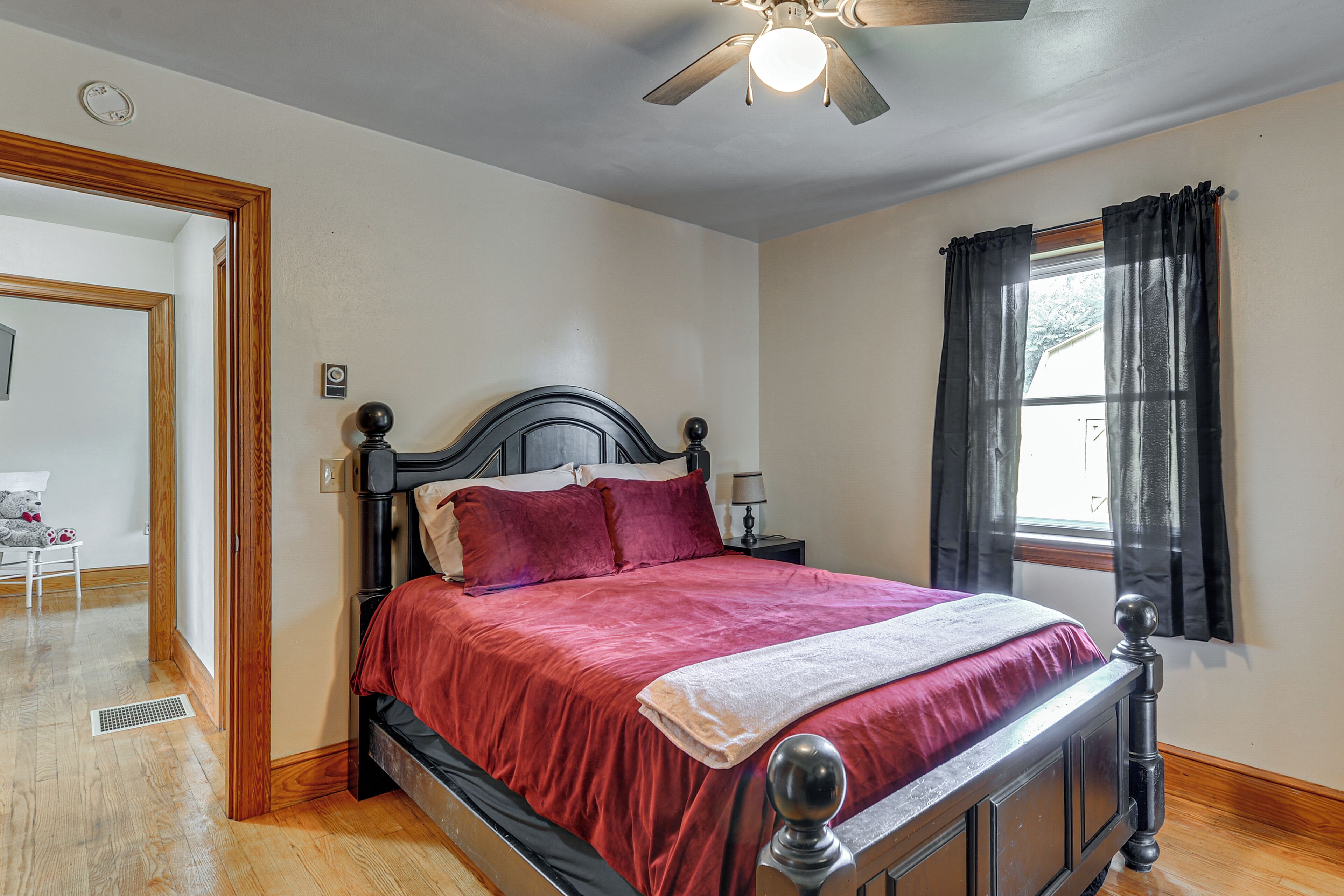 A bedroom with a bed, a ceiling fan, a window with curtains, and a door leading to another room.