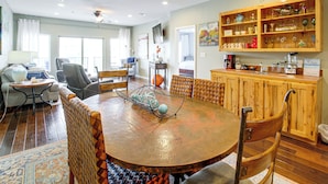 Amplia sala de estar con una mesa de comedor de madera rodeada de sillas, un área de cocina con armarios de madera y una zona de estar con sofá y sillones.