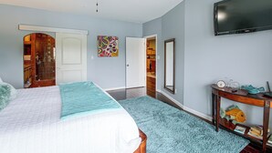 Un dormitorio con cama, ventilador de techo, televisor y puerta que da a un baño.