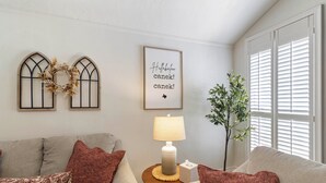 A living room with a grey sofa, a small table with a lamp, a wall-mounted picture, and a window with blinds. - The Oaks at Aggieland! Modern Home w/ Patio & Yard (Bryan)