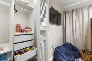 Um quarto infantil com uma estante branca, televisão e um espaço aconchegante para sentar com almofada azul e um cesto.