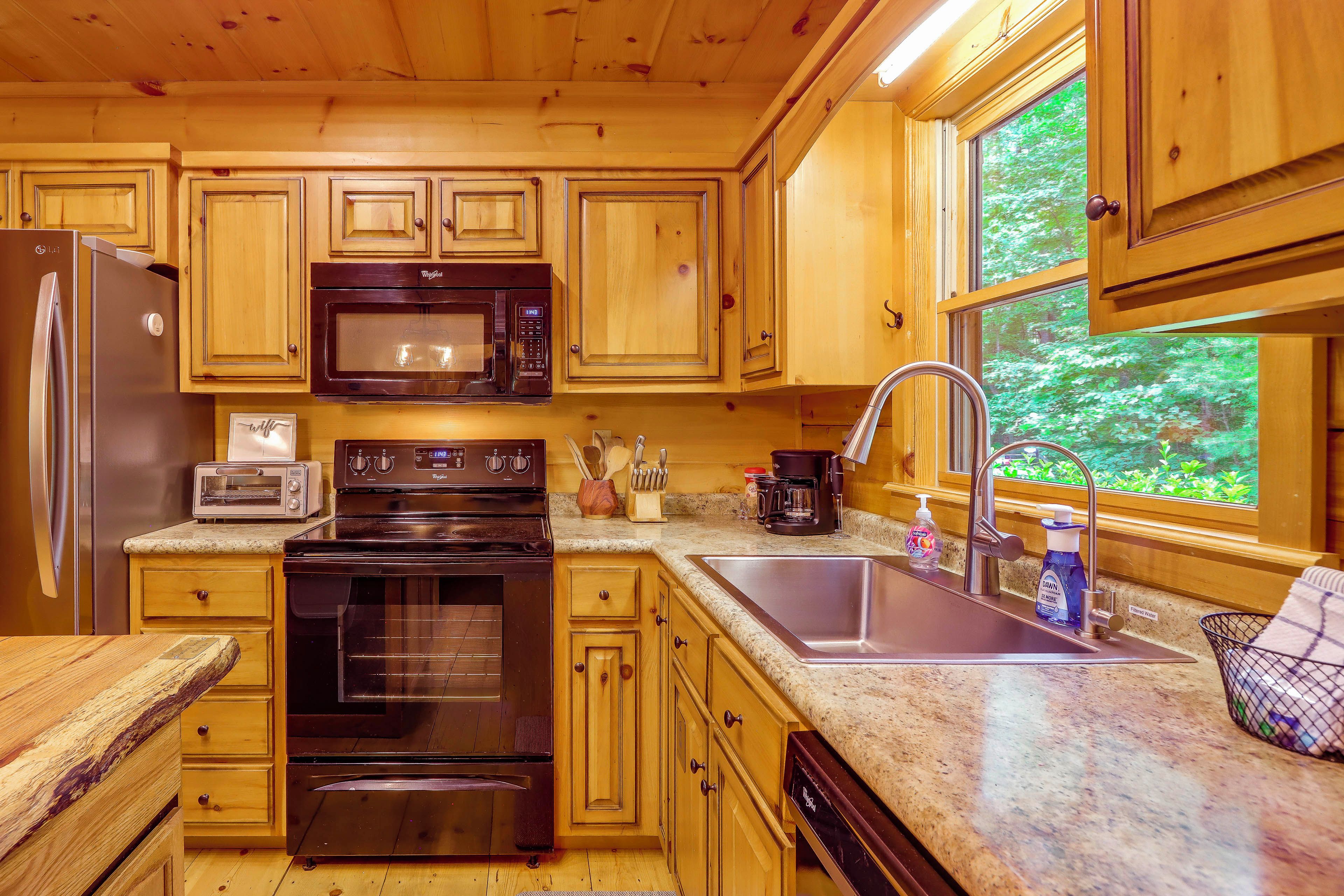 A kitchen with wooden cabinets, a microwave, a stove, a sink, and a window with a view of greenery.