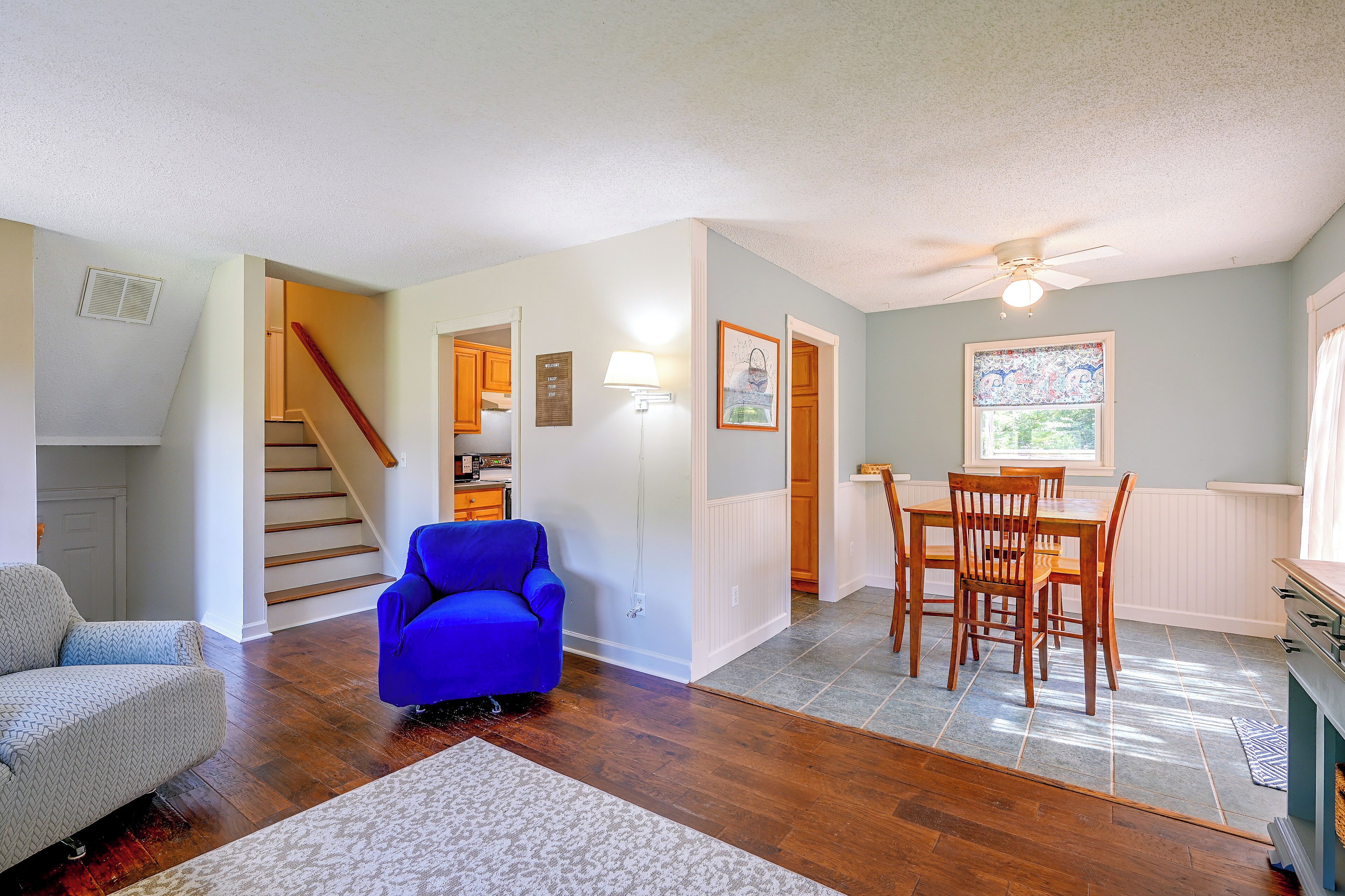 A spacious living area with hardwood flooring, a blue armchair, a dining table with chairs, and a staircase leading to another level.