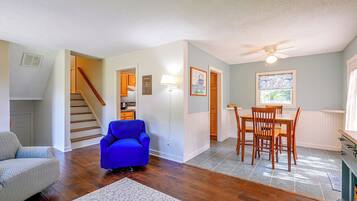 A spacious living area with hardwood flooring, a blue armchair, a dining table with chairs, and a staircase leading to another level.