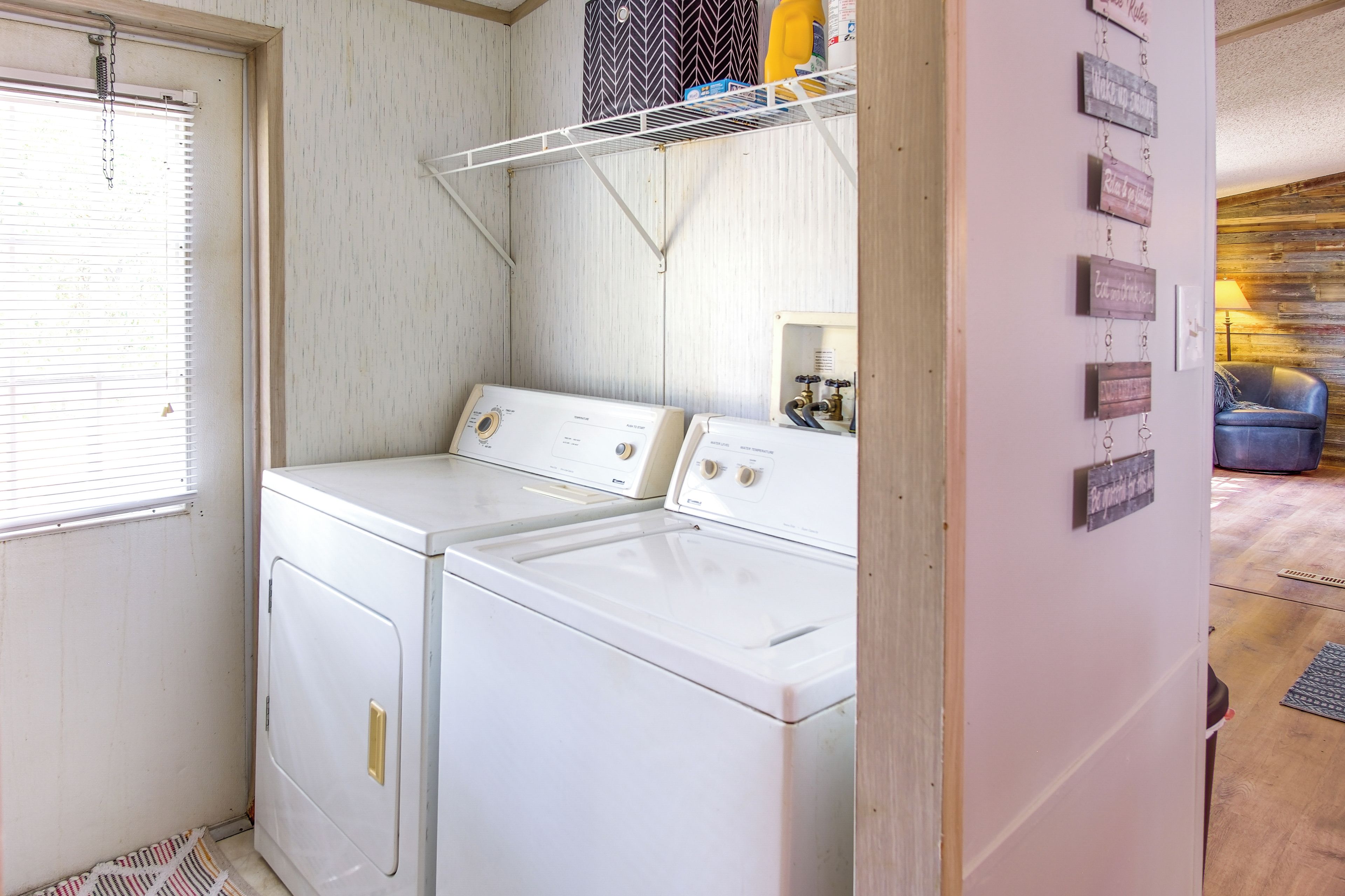 A laundry room with two white washing machines, a metal shelf above, and a window with blinds.