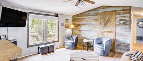 A cozy living room with a ceiling fan, a fireplace, a large window, and a rustic wooden wall.