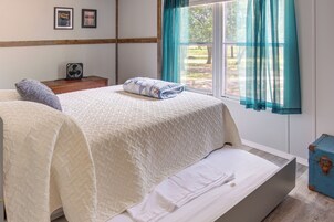 A bedroom with a bed, a bedside table with a fan, a window with curtains, and a suitcase.